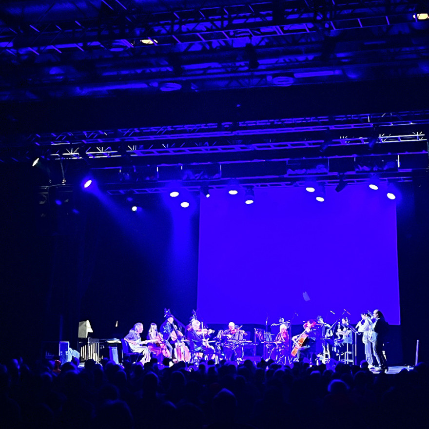 Kronos Quartet and Summer Festival Musicians plus All-Stars perform onstage in blue lighting at MASS MoCA's main auditorium.